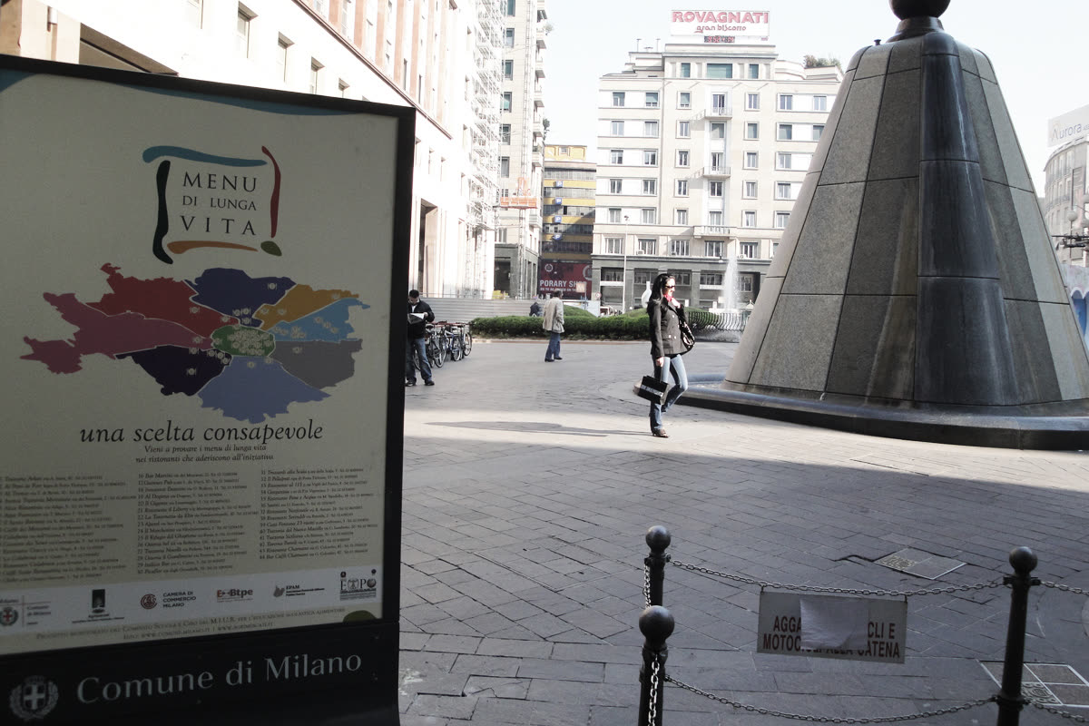Totem stradale Menu di Lunga Vita in una piazza di Milano con passanti e fontana