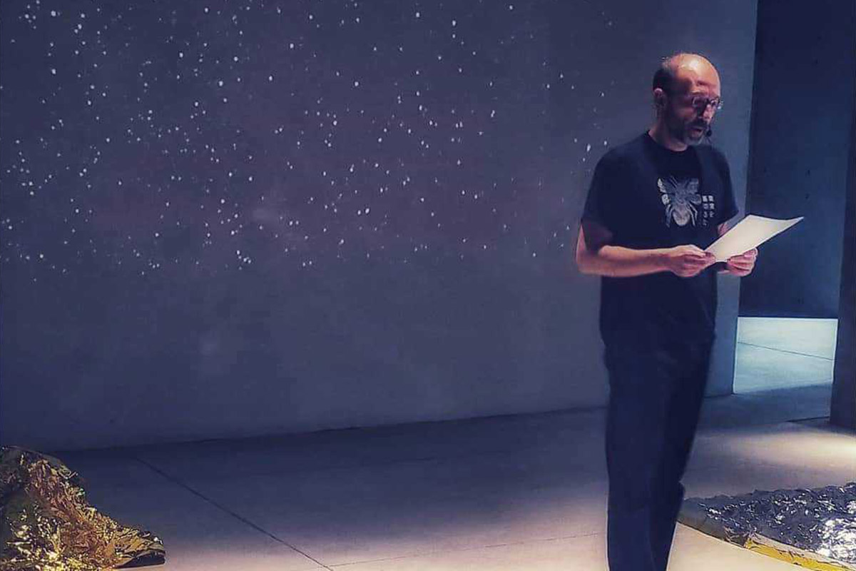 Mauro Covacich con microfono ad archetto legge davanti alla parete di luci stellate