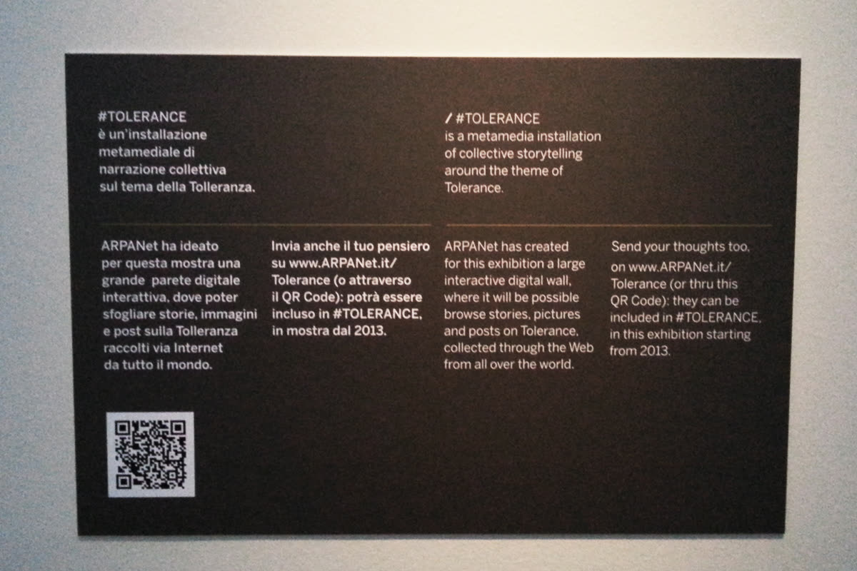 Pannello espositivo bilingue di #TOLERANCE con testo introduttivo e QR code su parete